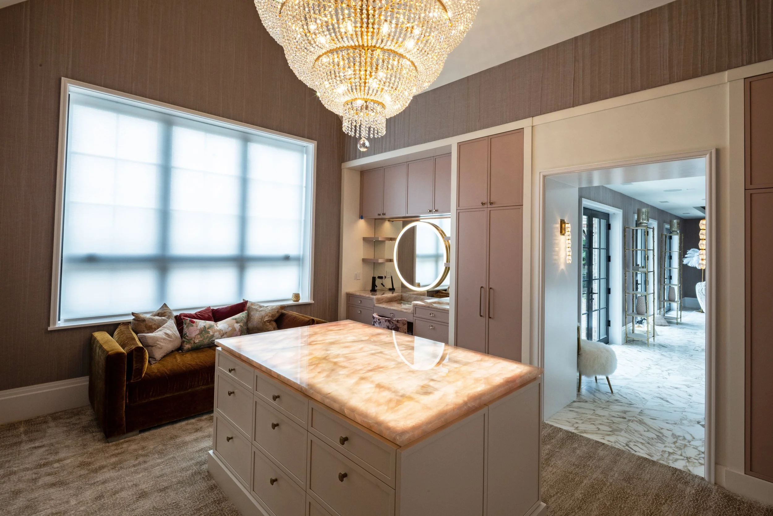 Interior view of a modern home with a large, elegant chandelier, a window with white blinds, a cozy seating area with pillows, a marble-topped island, and a door leading to another room with gold and glass decor.
