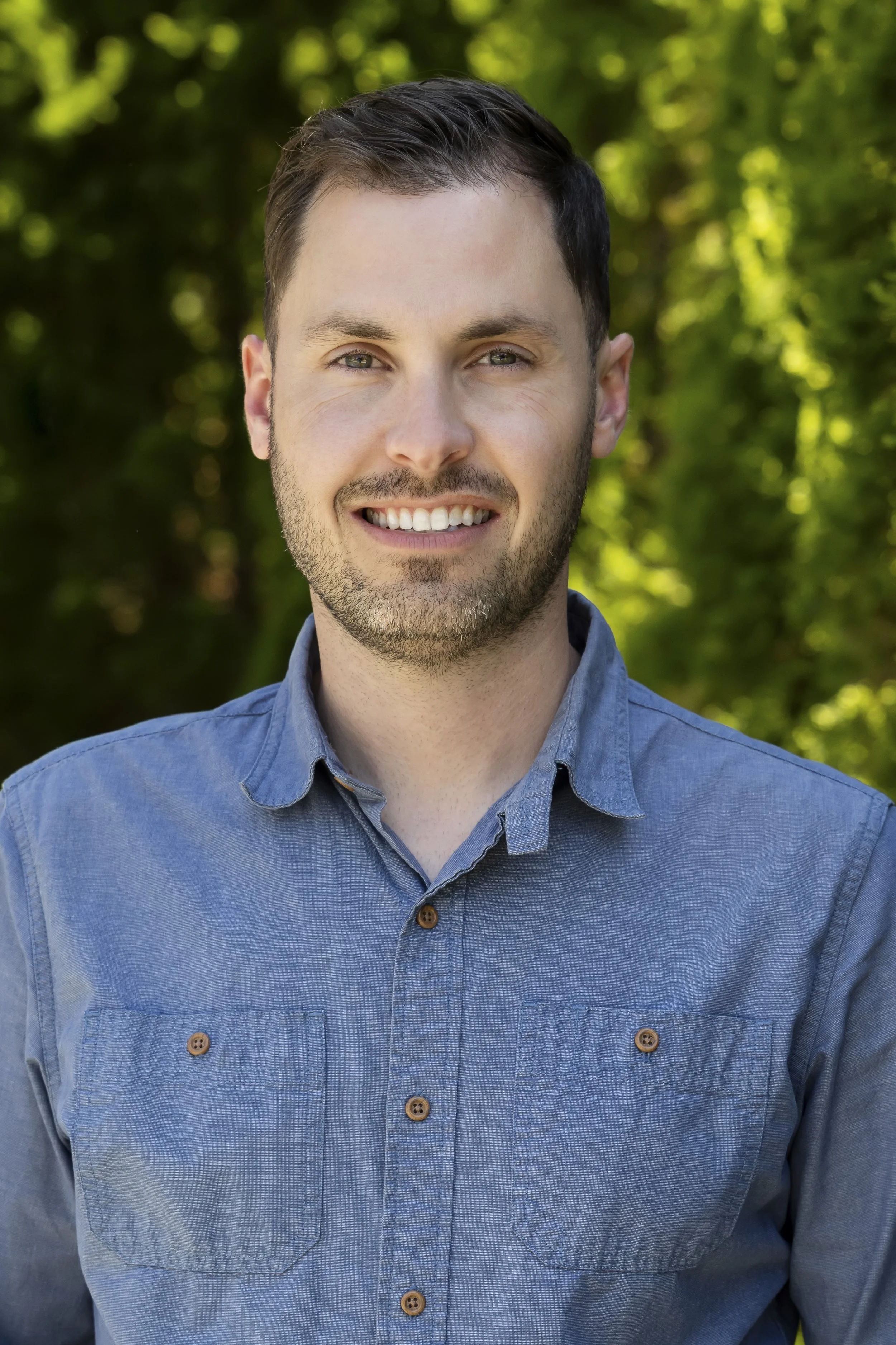 A man with short dark hair, slight facial hair, and green eyes, smiling while wearing a blue button-up shirt. He is outdoors with a blurred green leafy background.