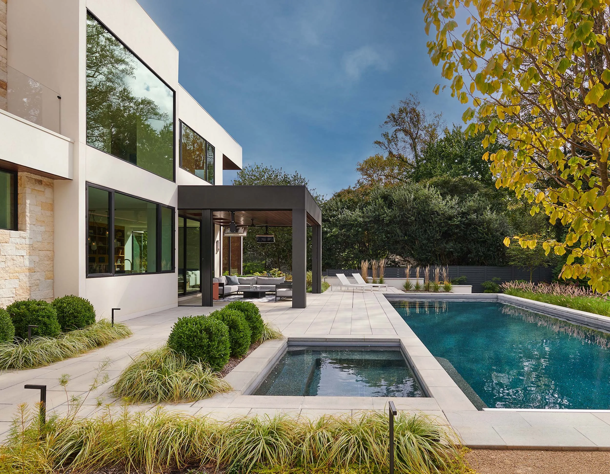 Modern house with large glass windows, outdoor patio with seating, swimming pool, and landscaped yard with trees and bushes under a blue sky.