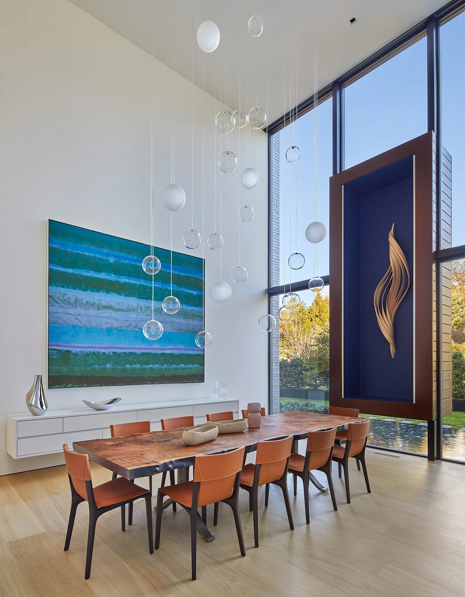 Modern dining room with a large wooden table, brown chairs, a white sideboard with decorative vases, a colorful abstract painting on the wall, and a large window with a view outside. Decorative glass and white orbs hanging from the ceiling.