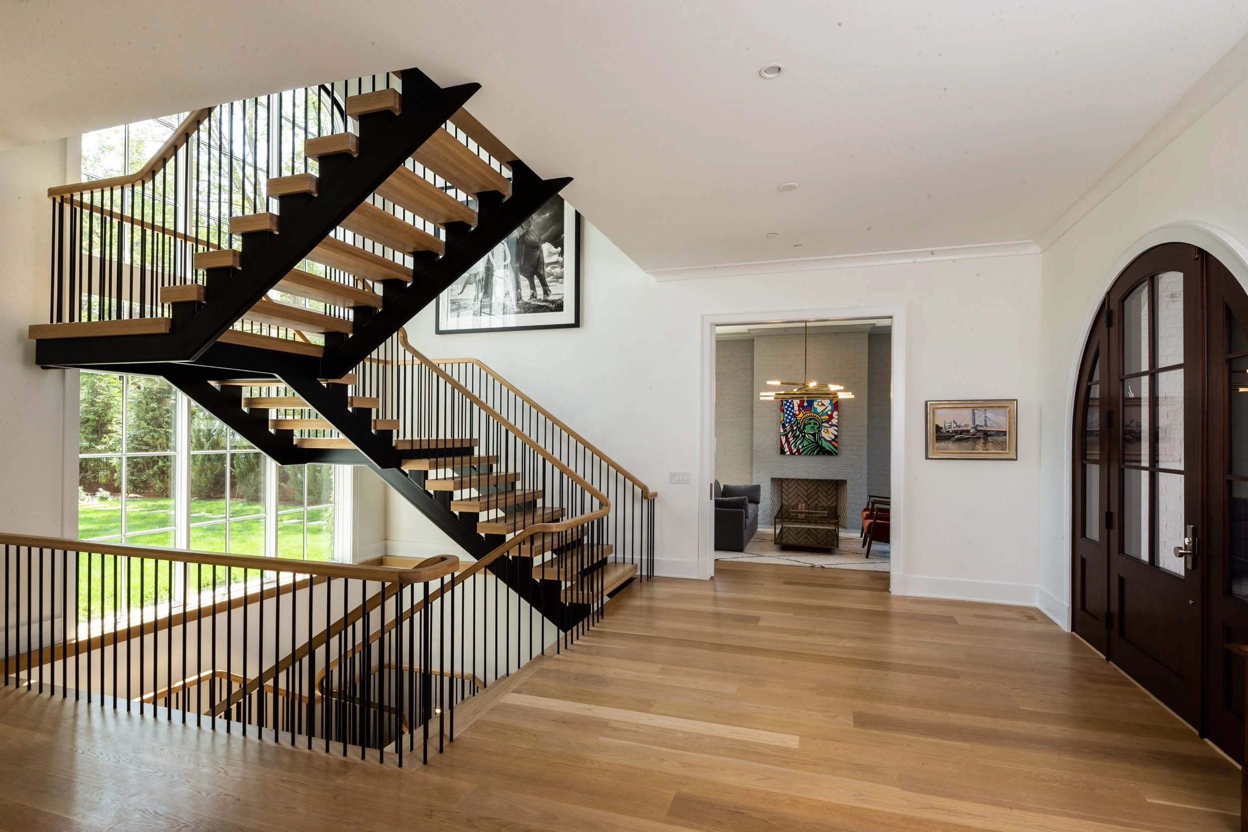 Modern home interior with staircase, large windows, artwork, and a living area visible through an open doorway.