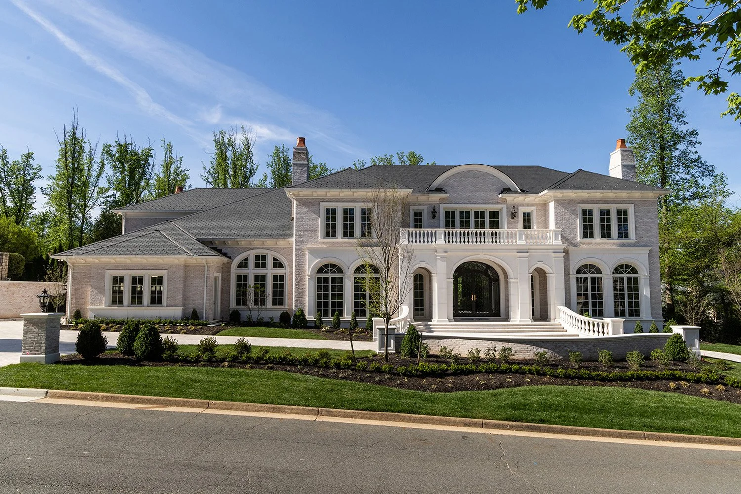 A large, elegant house with a white exterior, multiple large windows, and a front porch with a staircase. The house is surrounded by well-maintained landscaping, including grass, shrubs, and young trees, under a clear blue sky.