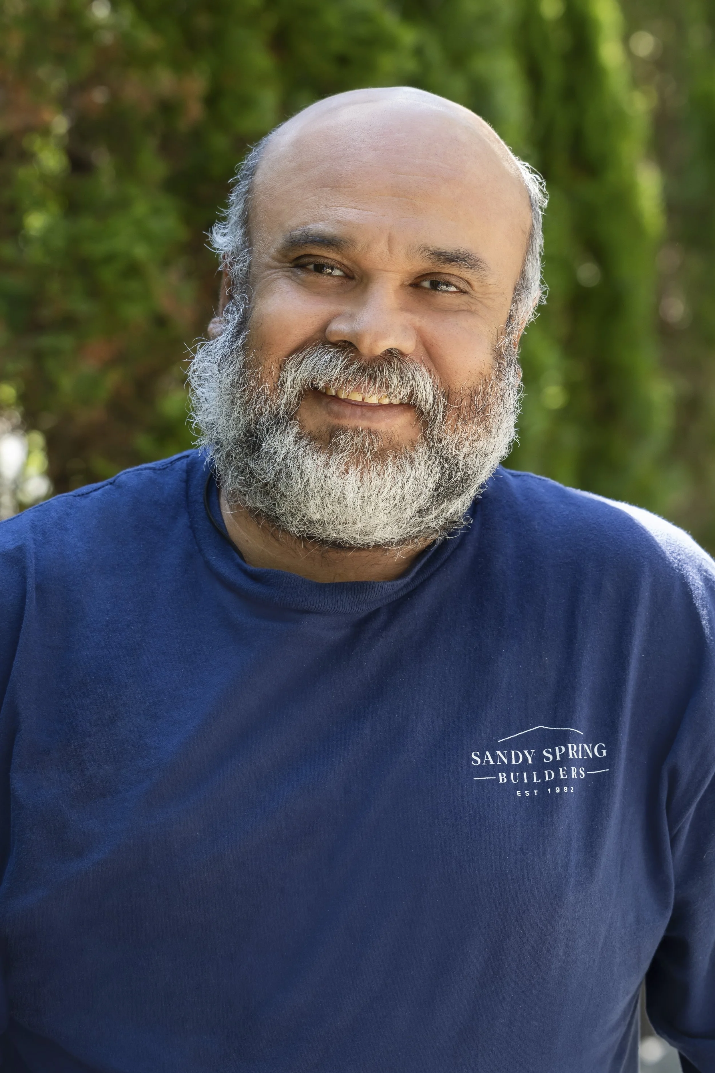 A smiling man with a beard and mustache wearing a navy blue shirt with the logo 'Sandy Spring Builders Est 1982' on the chest, outdoors with greenery background.