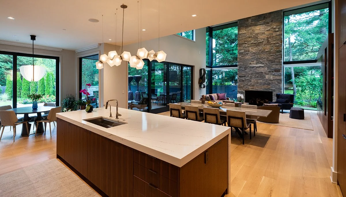 Open-concept kitchen and living room with large windows and modern decor, including a kitchen island, dining table, and sitting area with fireplace.