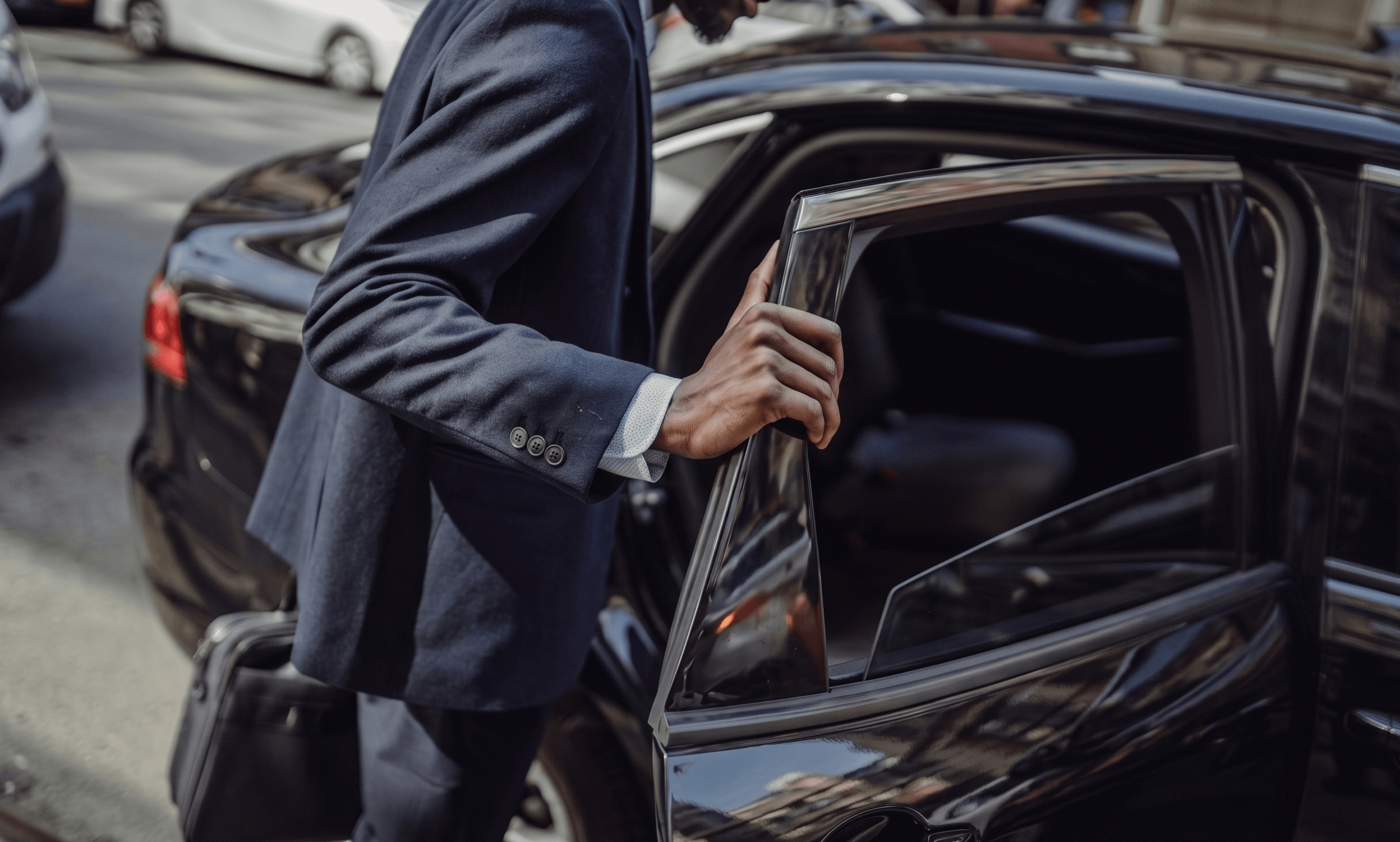 High-definition close-up of a man in a navy blue suit with a briefcase entering the back seat of a glossy black executive sedan on a city street.