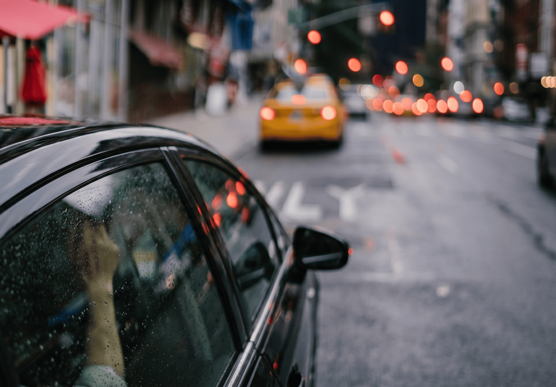 A city street scene with a car in the foreground, rain droplets on the window, and blurred cars and city lights in the background
