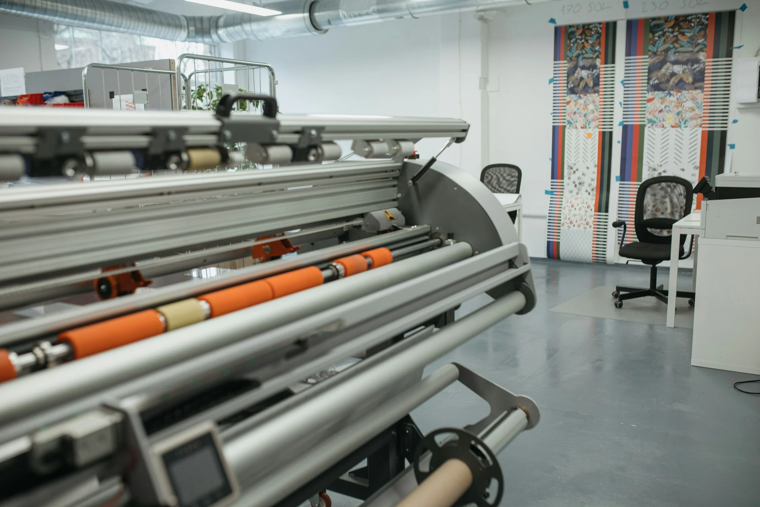 An industrial workspace with a large printing or plotting machine in the foreground and two office chairs, a desk, and colorful patterned wall hangings in the background.