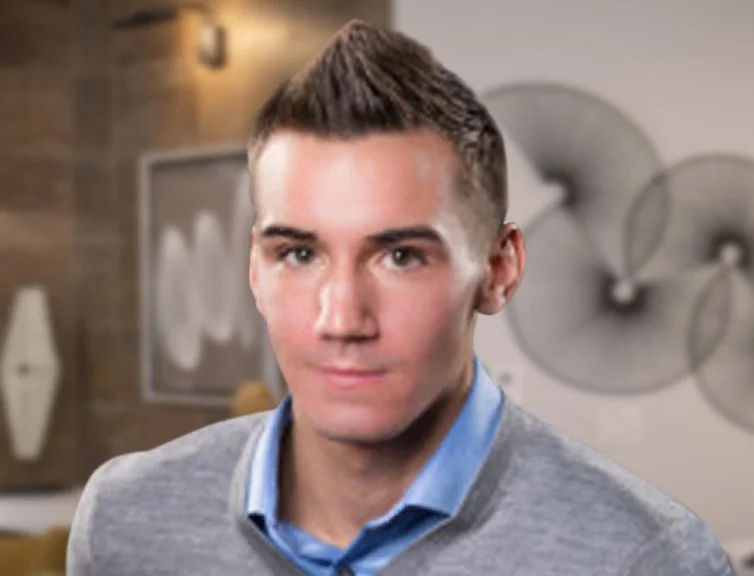 A young man with short, styled brown hair wearing a light blue collared shirt and a gray sweater in a modern decor setting.