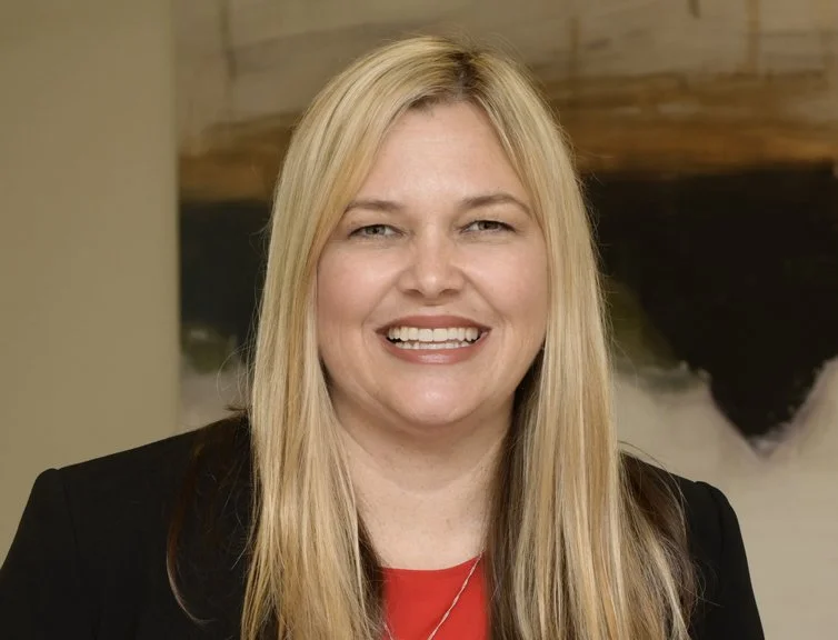 A woman with long blonde hair smiling, wearing a black blazer and red top, standing indoors.