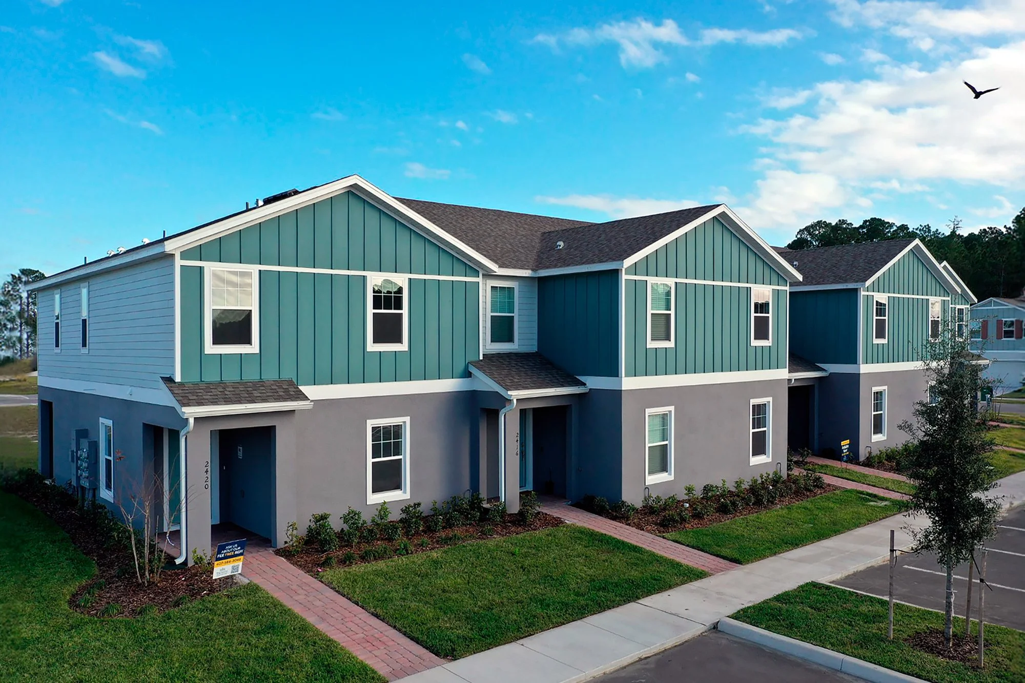 Two-story modern townhouses with teal and gray exteriors, small front lawns, brick walkways, and a parking lot. Blue sky with some clouds and a bird flying overhead.