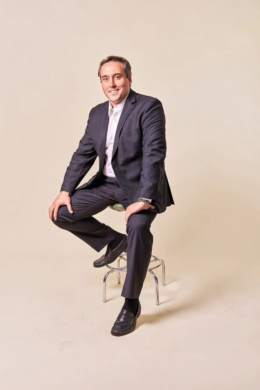 A man in a dark suit and white shirt sitting on a stool against a plain light-colored background.