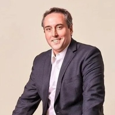 A man in a dark suit smiling, with a neutral background.