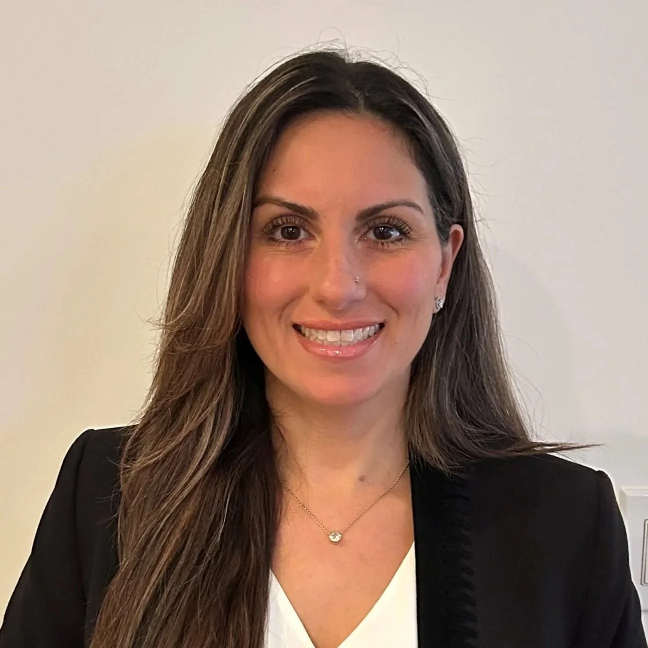 Portrait of a woman with long brown hair wearing a black blazer, white top, and a delicate necklace, smiling against a plain light-colored background.