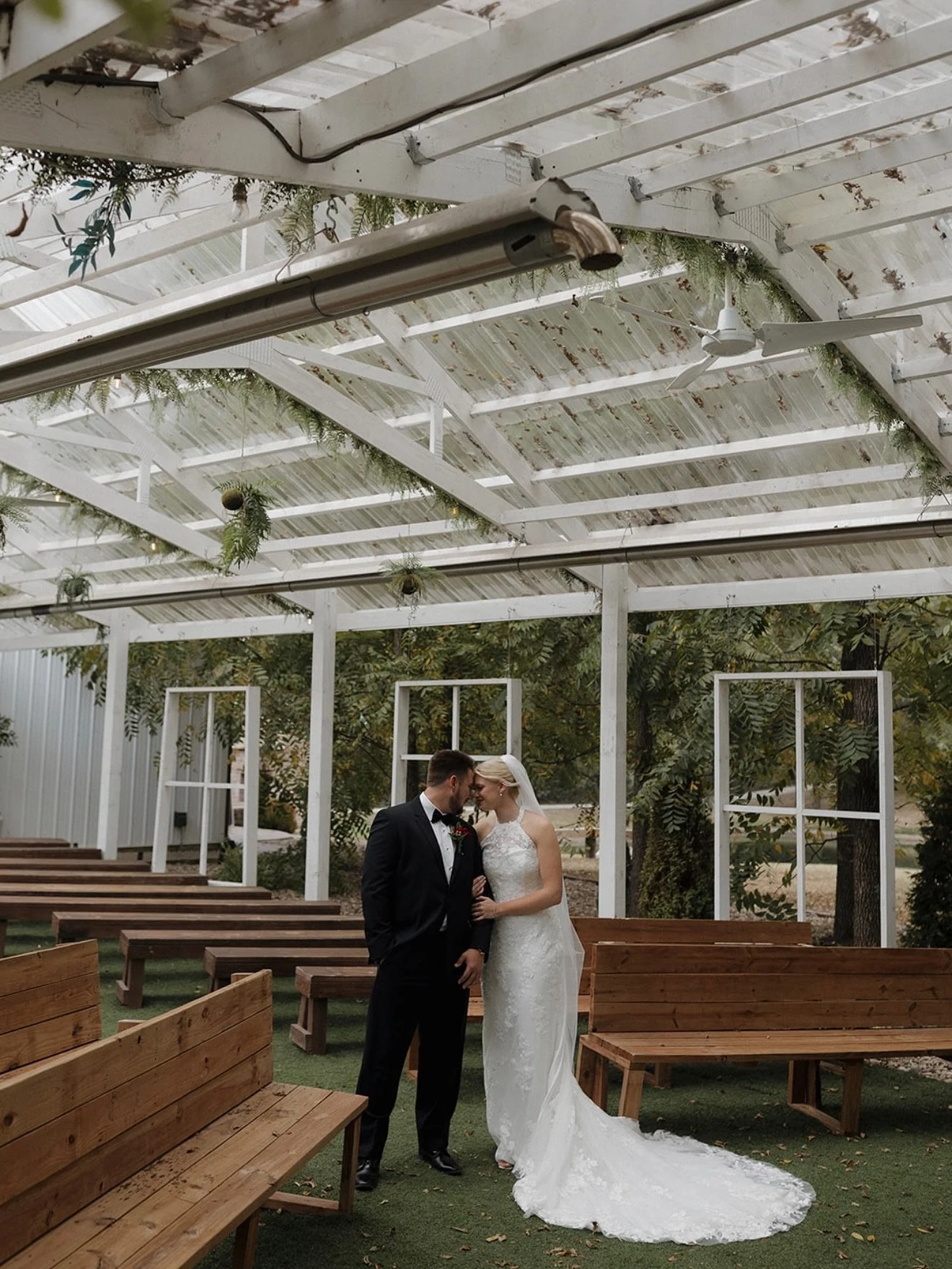 A bride and groom dressed in wedding attire face each other inside an outdoor wedding venue with wooden benches, white lattice structures, and trees in the background.