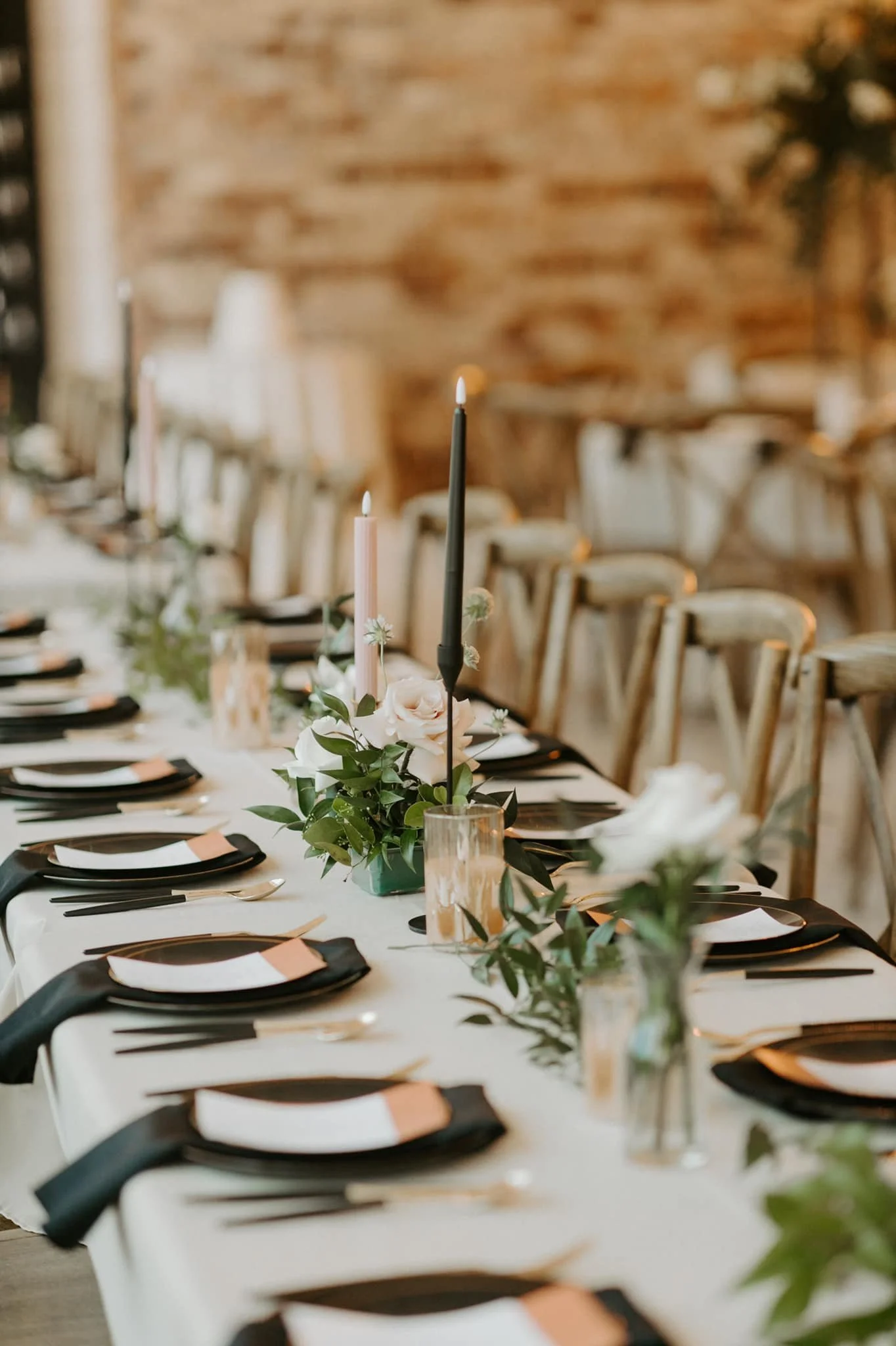A long dining table set for a special occasion with black napkins, gold flatware, and white plates, decorated with pink candles and floral centerpieces, in a rustic setting with wooden chairs and a brick wall.