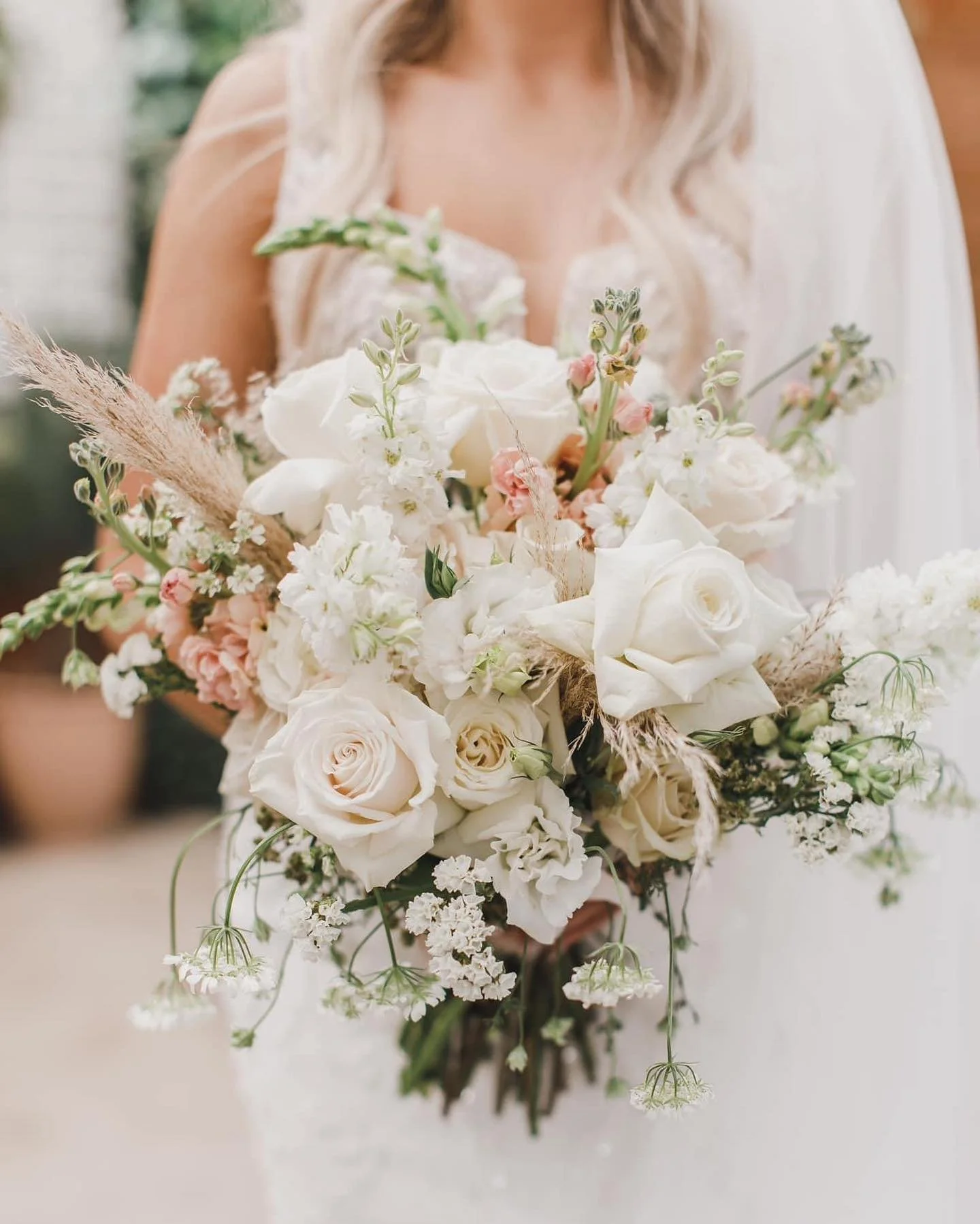 A bride holding a large bouquet of white and pale pink flowers, including roses, with greenery and dried grasses, in an outdoor setting.