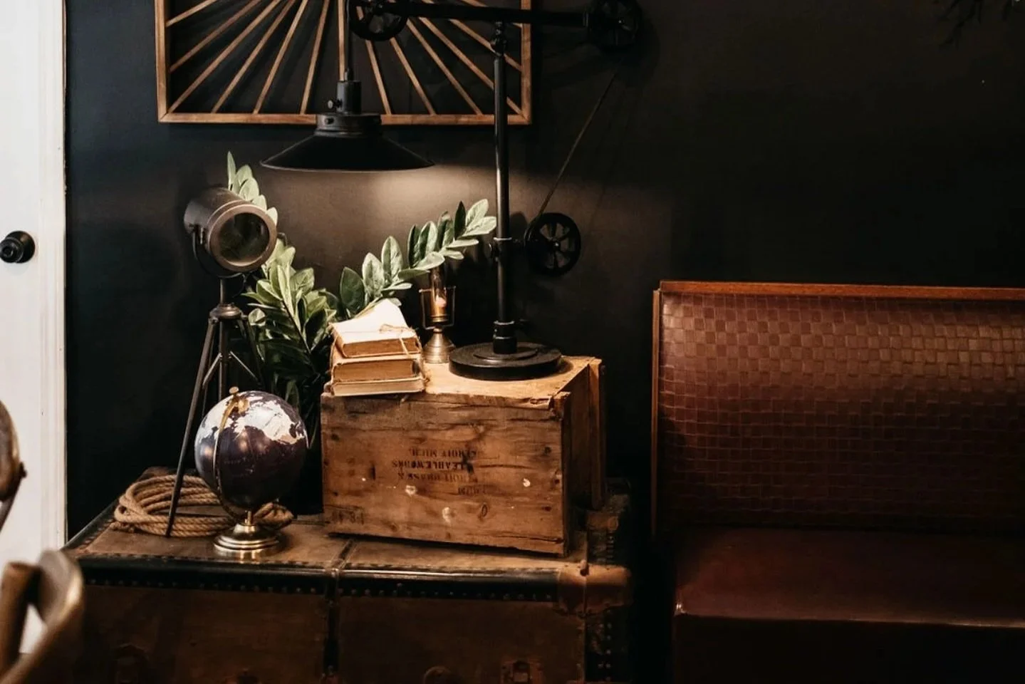 Decorative vintage-style room corner with lantern, globe, stack of books, green plants, and wooden crates on a wooden table, against a black wall with a framed geometric wood art piece.