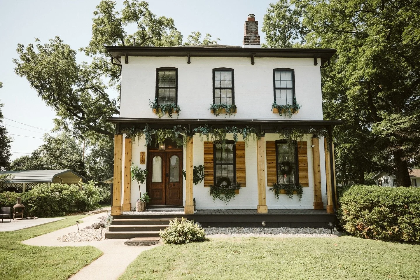A two-story white house with black window frames and shutters, a brick chimney, and a front porch supported by wooden columns. The house is decorated with greenery and flower boxes, with stairs leading up to the main door, and surrounded by a well-maintained lawn and trees.