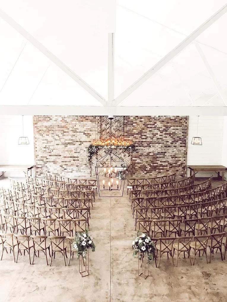 Indoor wedding ceremony setup with rows of wooden chairs facing a brick wall and a lit chandelier, decorated with floral arrangements at the aisle.