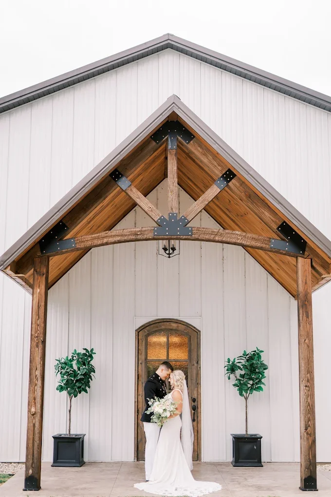 A bride and groom standing close, holding a bouquet, in front of a rustic barn with a wooden arch and two potted plants.