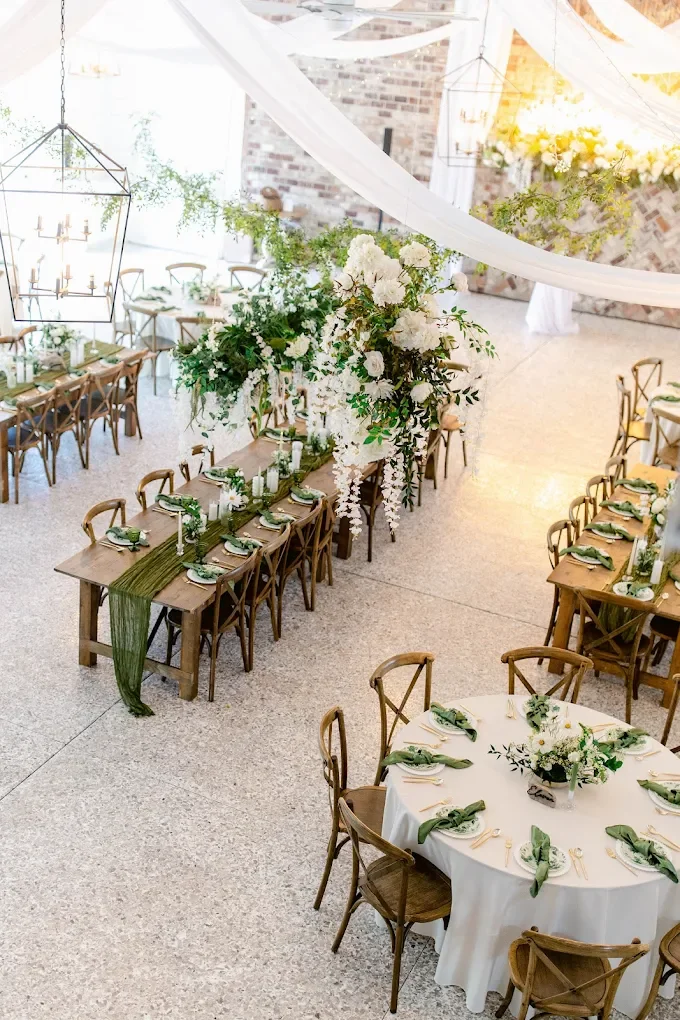 Elegant indoor wedding reception with wooden tables, white floral centerpieces, and draped fabric decor.