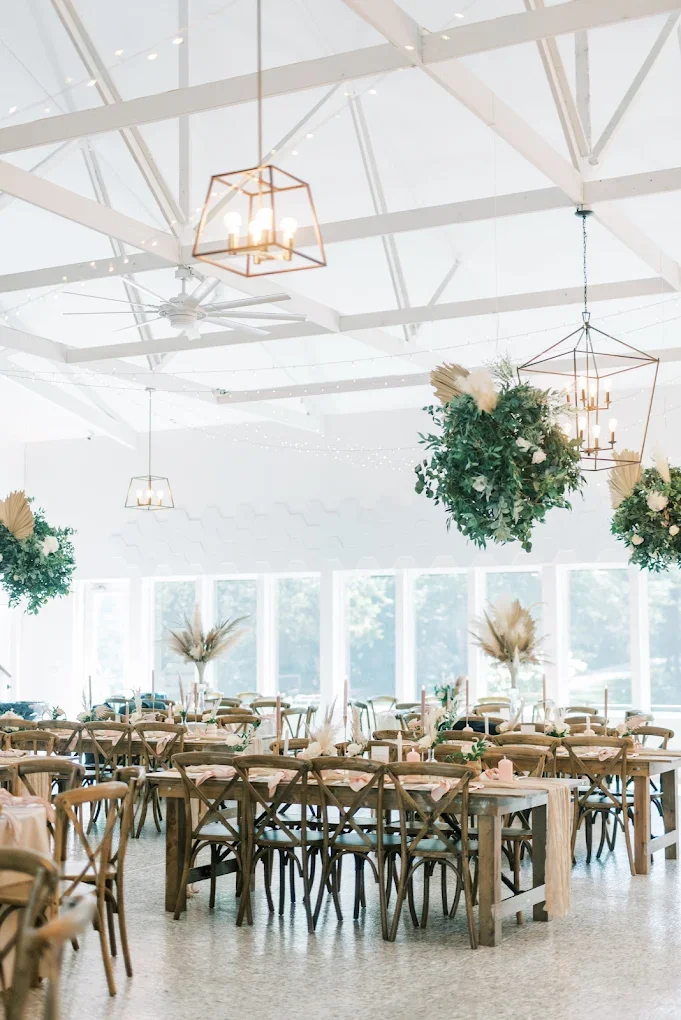 Bright event space decorated with hanging greenery and floral arrangements, wooden tables and chairs, large windows, and geometric pendant lights.