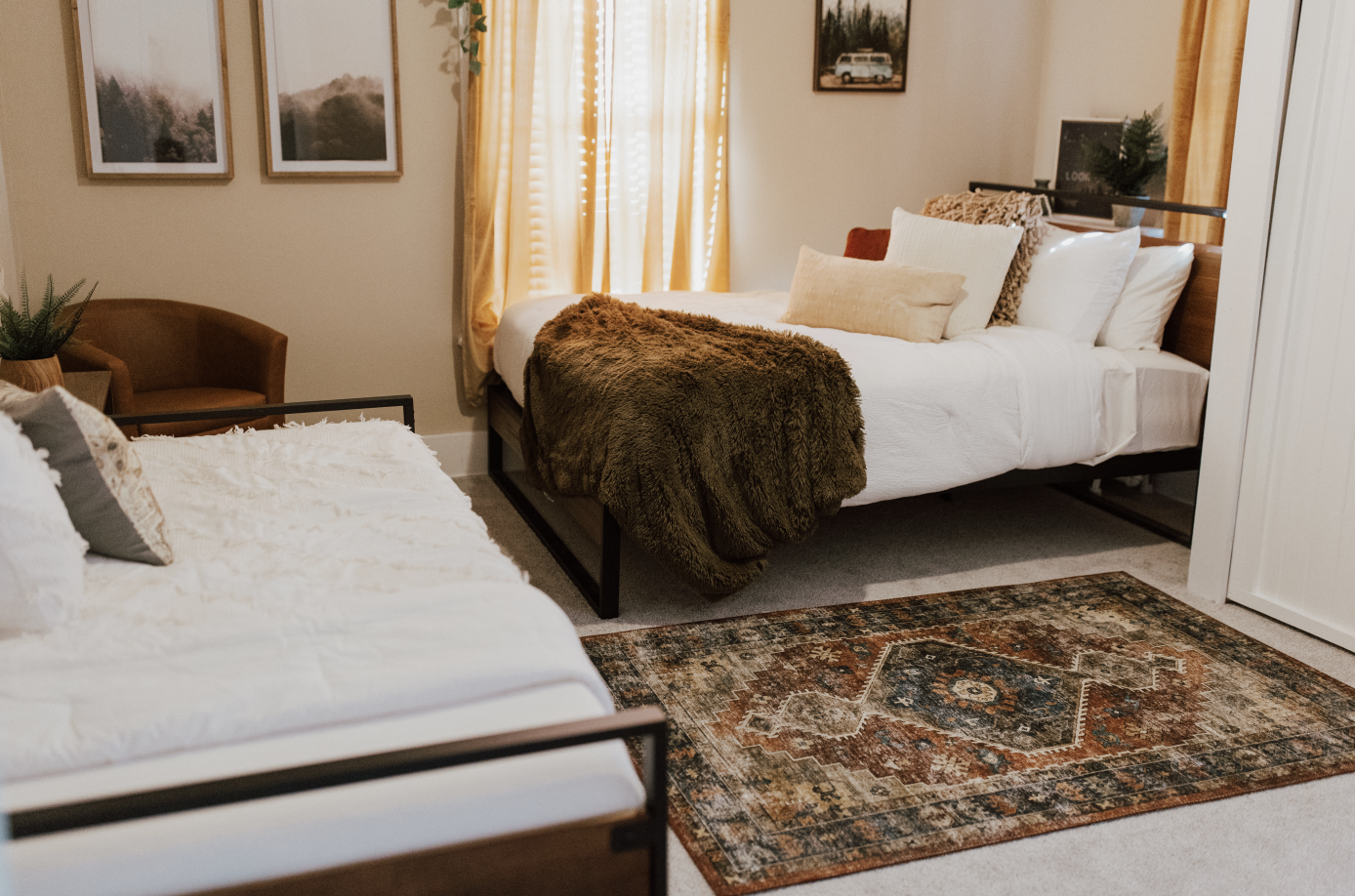 A bedroom with two beds, one with a brown furry blanket, pillows, and a rug. There is a window with yellow curtains, framed pictures on the wall, and a small chair with a plant next to it.