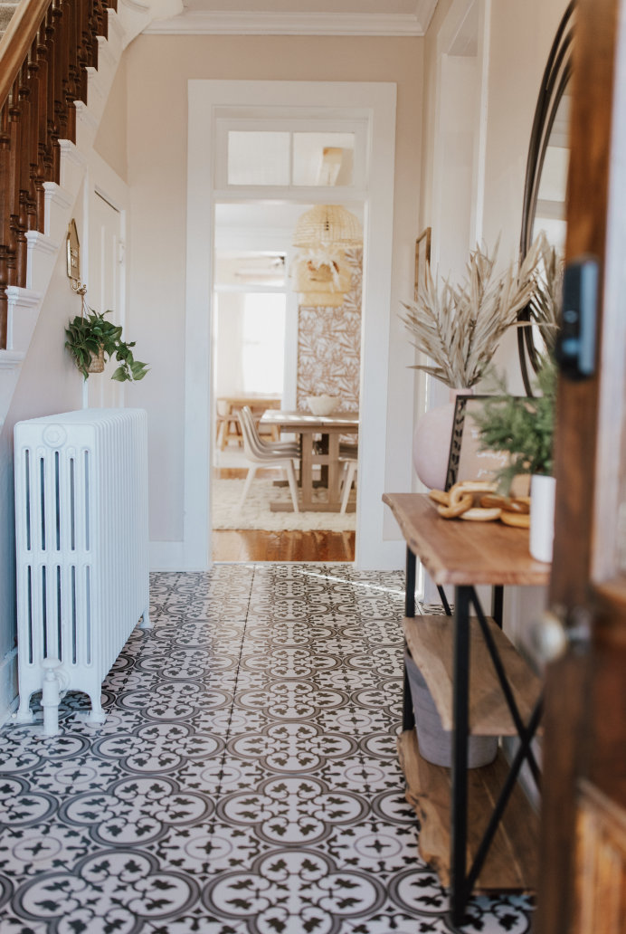 Hallway leading to a dining room with a wooden table and chairs, decorative patterned tiles on the floor, and a wooden shelf with plants and decorations on the right.