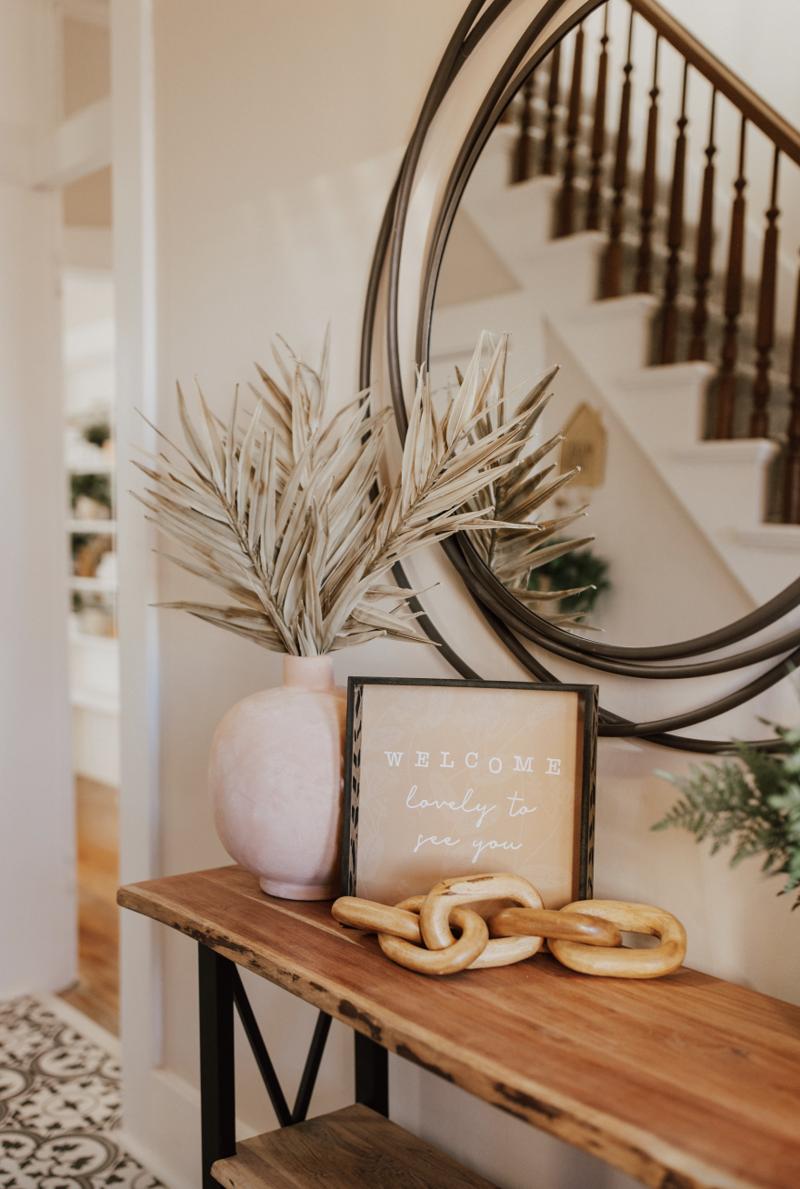 Decorative entryway table with a large pink vase holding dried palm leaves, a framed sign that reads 'Welcome, lovely to see you,' and a chunky wooden chain, with a round mirror reflecting a staircase in the background.