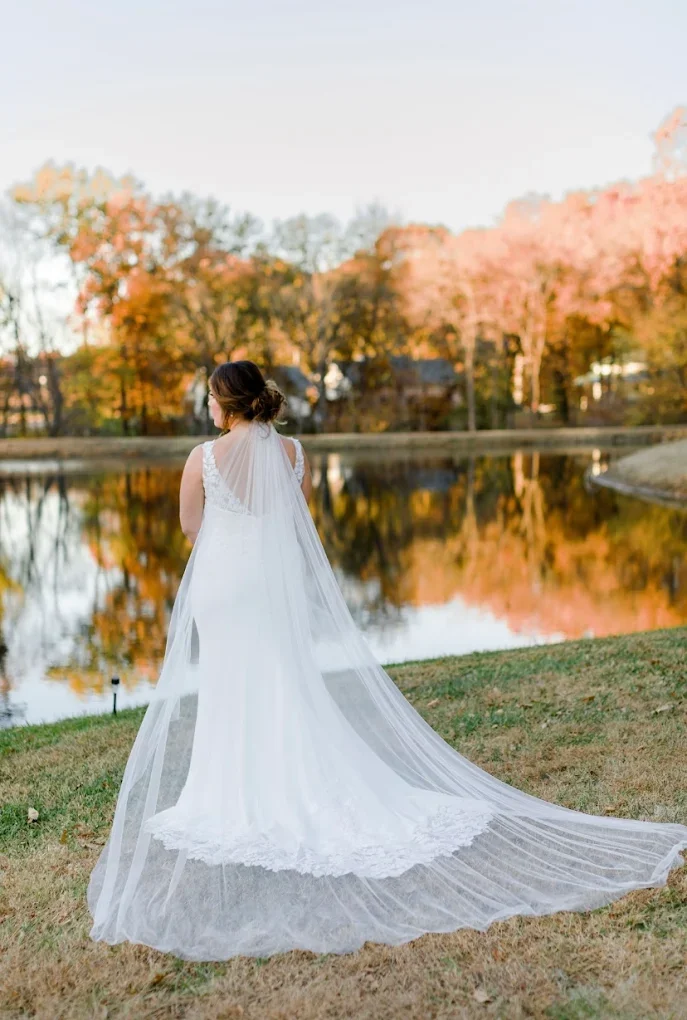 A bride in a white wedding dress with a veil, standing near a pond with colorful autumn trees reflected in the water.