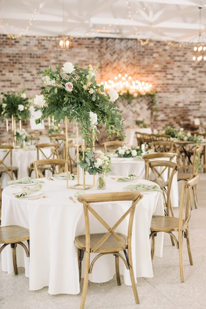 Wedding reception setup with round tables, white tablecloths, floral centerpieces, and wooden chairs in a decorated venue.