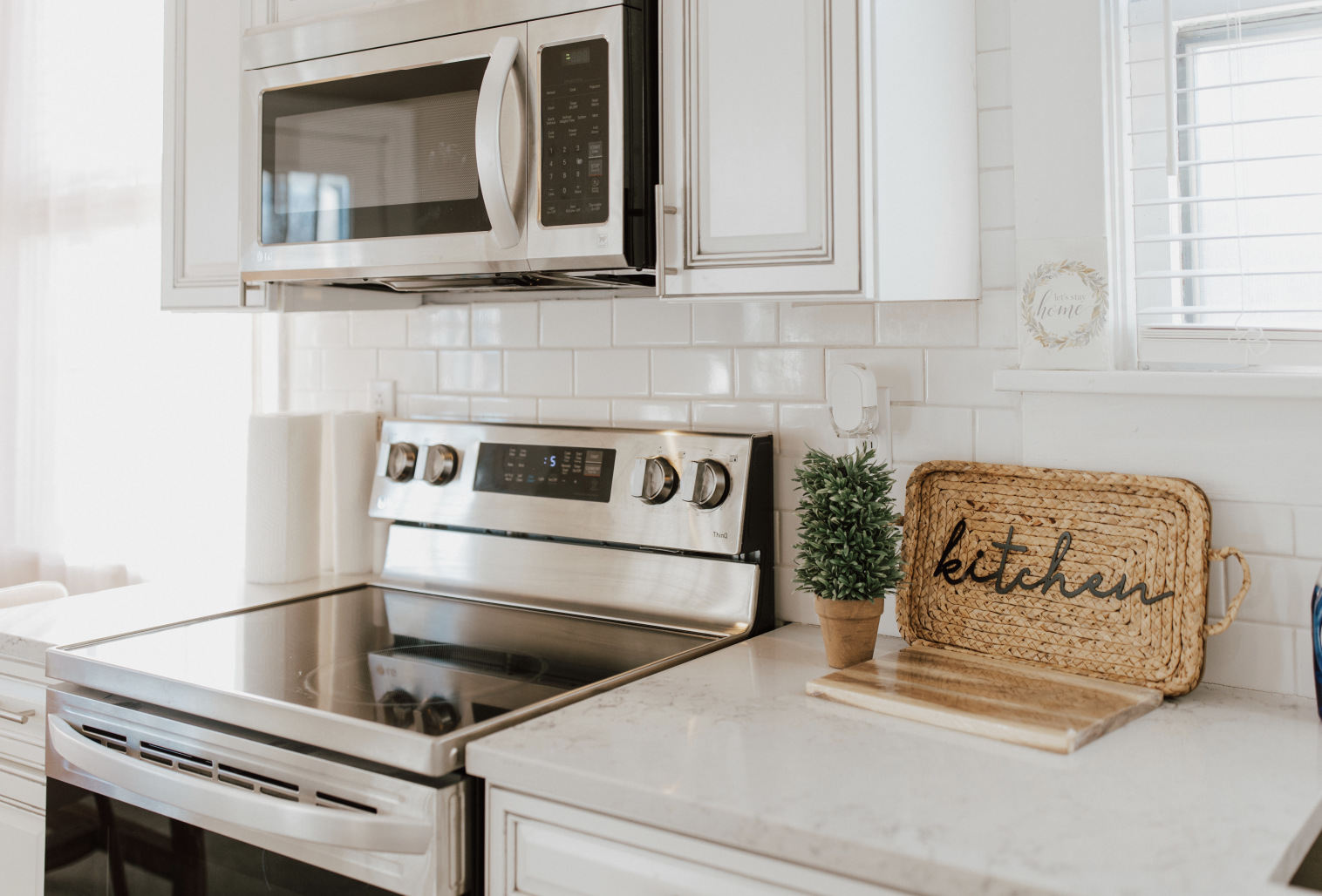 Modern kitchen with white cabinets, stainless steel microwave and stove, decorative plant, wooden cutting board, and a woven tray with the word 'kitchen' on it, near a window with white blinds.