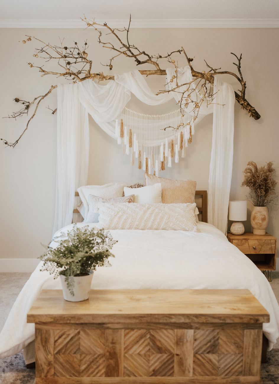 A bedroom decor with a carved wooden bench at the foot of the bed, featuring a small potted plant. The bed has multiple cream and white pillows, a white bedspread, and is framed by white drapes and a large branch with hanging tassels above the headboard. A nightstand with a lamp, a ceramic vase with dried plants, and a decorative pot is beside the bed.