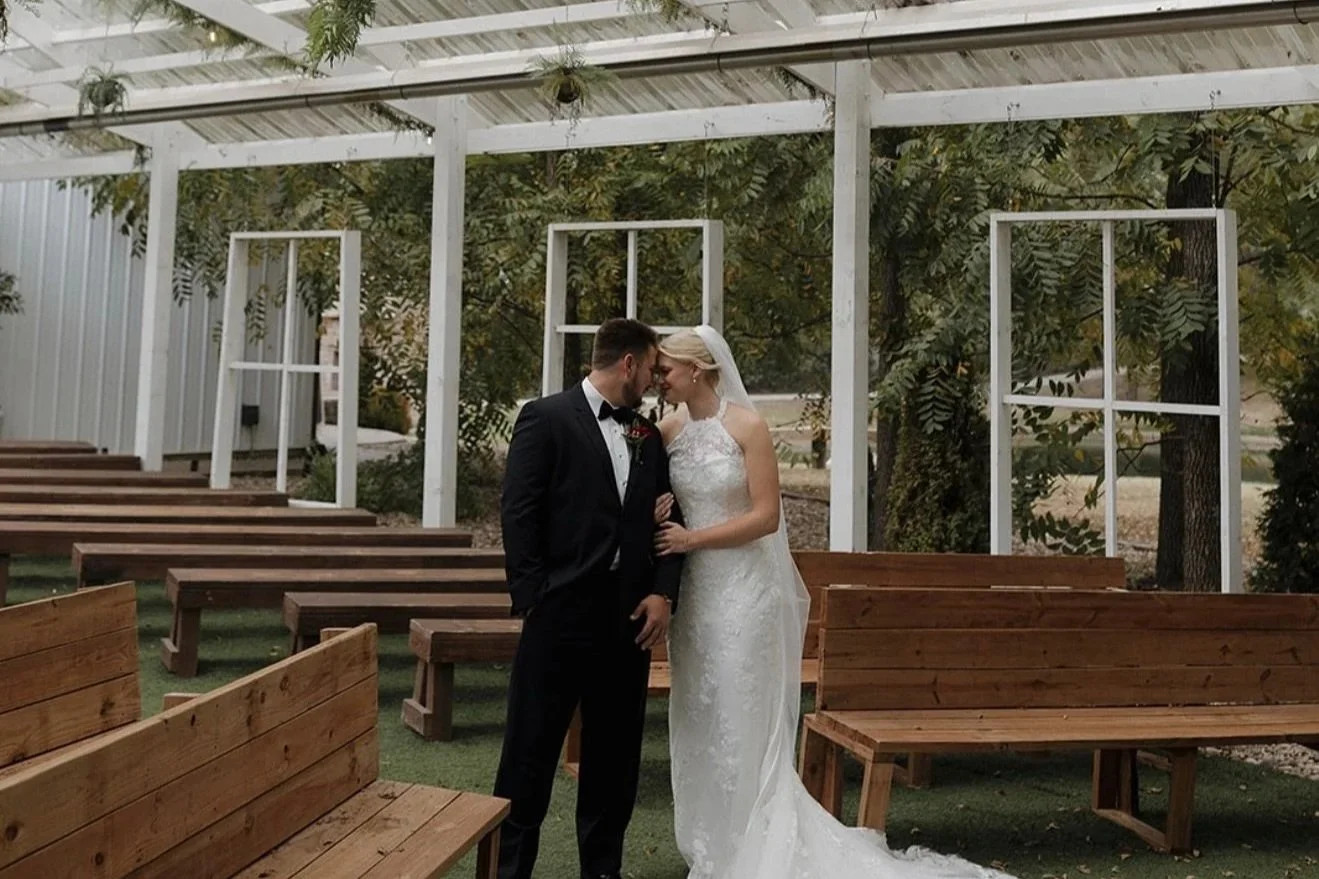 A bride and groom dressed in wedding attire standing close together in an outdoor setting with benches and a white framework, touching foreheads.