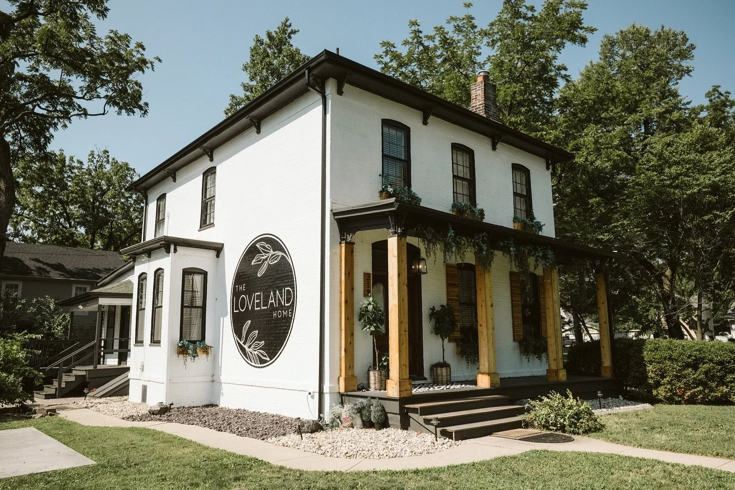 A white two-story building with black trim and a sign that reads 'The Loveland Home'. The building has a wooden front porch with steps, potted plants, and greenery hanging from the porch. There are trees and a sidewalk in front.