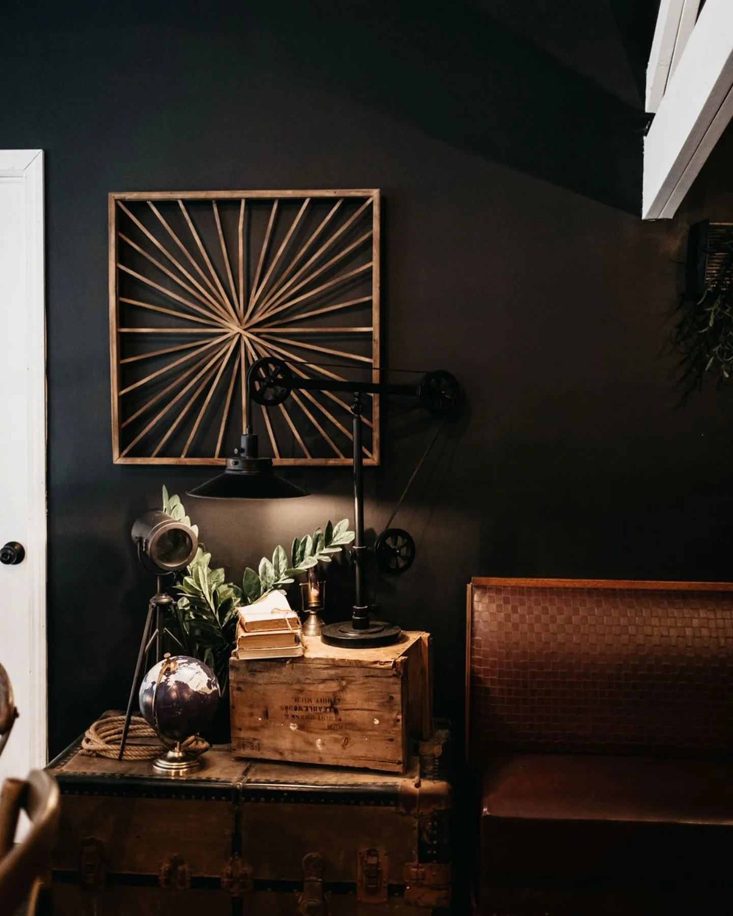 Vintage industrial decor with a black wall, wooden crates, an old globe, a plant, a lamp, and a geometric wooden wall art piece.