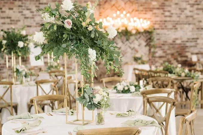 A wedding reception setup with floral centerpieces on round tables, gold chairs, and soft lighting in an indoor venue with brick walls.