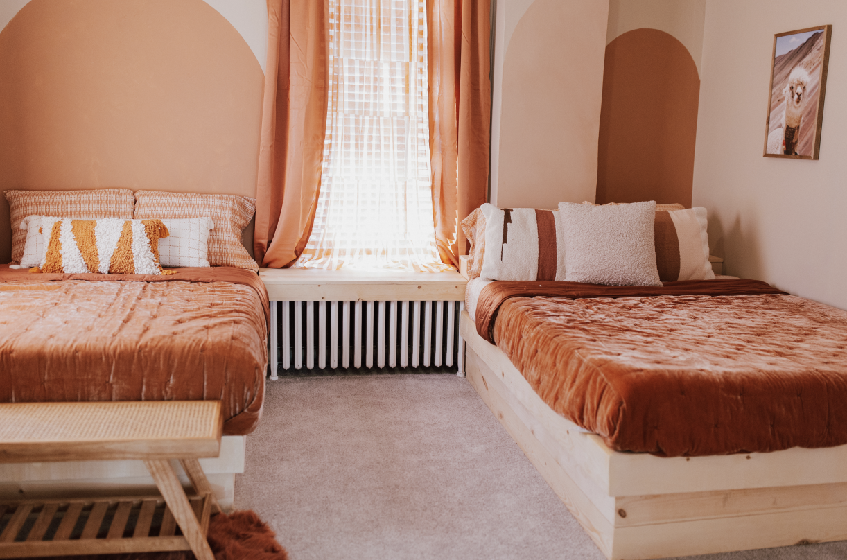 A cozy bedroom with two beds featuring rust-colored bedsheets, decorative pillows, a window with orange curtains, and a framed llama picture on the wall.