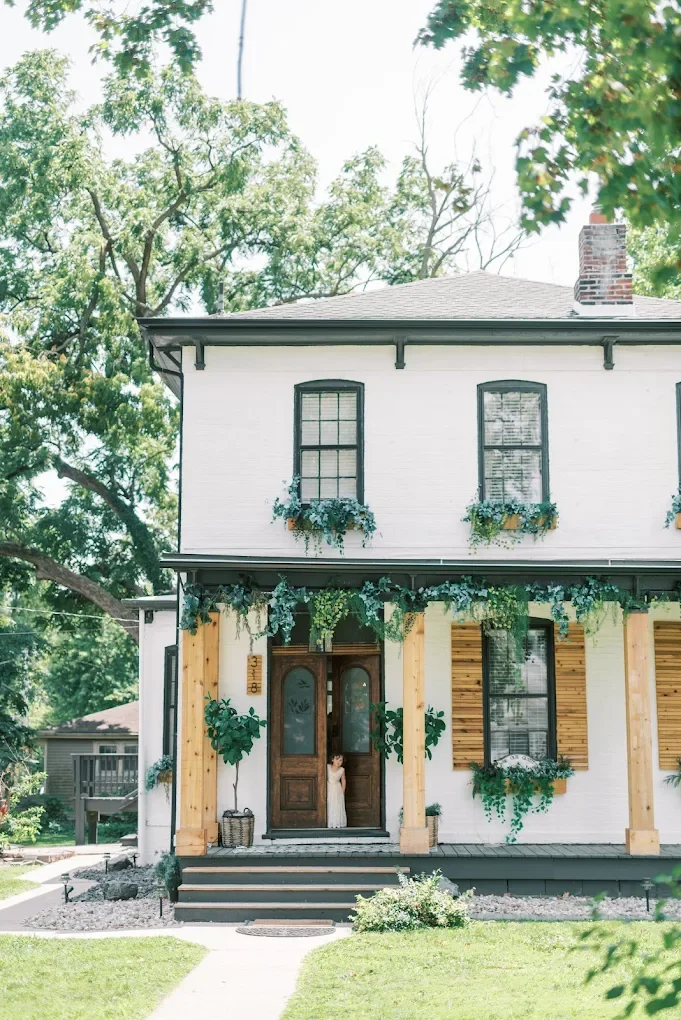 A two-story house with a white brick exterior, black window frames, and a chimney. The front porch is decorated with greenery and potted plants. A young girl is standing at the front door, which has a wooden finish and arched glass panels.