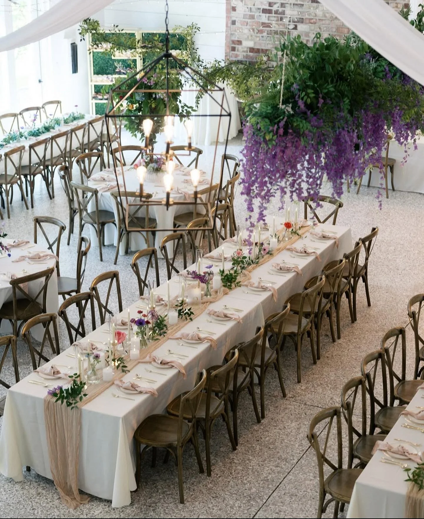 Wedding reception setup with long rectangular tables covered in white tablecloths, decorated with pink napkins, candles, and floral arrangements. Hanging purple and green floral decorations are suspended from the ceiling. Wooden chairs surround the t