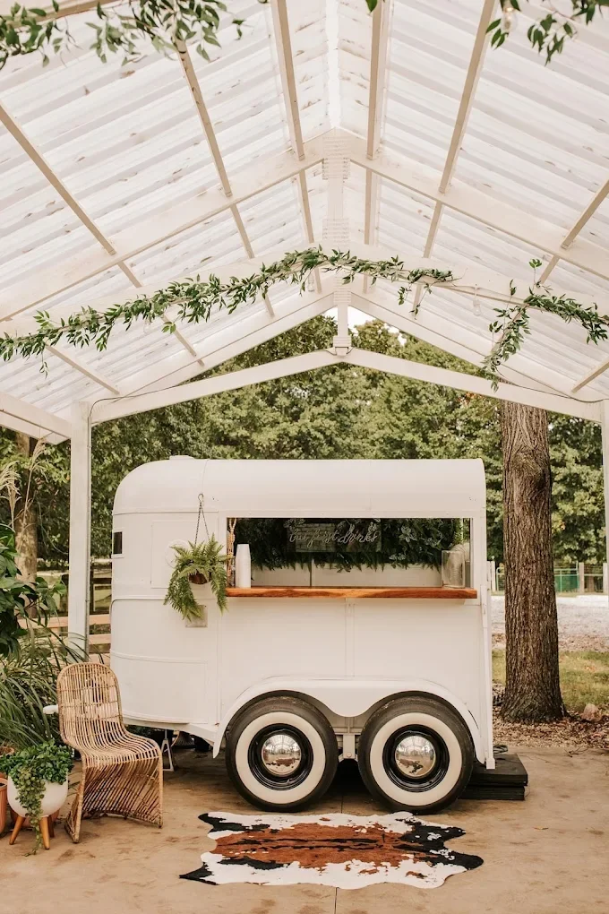 A white vintage food truck mounted on a trailer under a white metal shed decorated with green foliage, with a rattan chair, potted plants, and a cowhide rug in front.