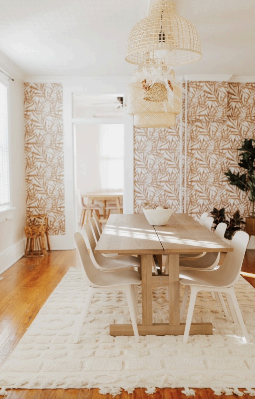 A dining room with a wooden table, six white chairs, a white bowl on the table, and patterned beige and white wallpaper. There are bright windows, a woven pendant light, a basket, and a small plant in the room.