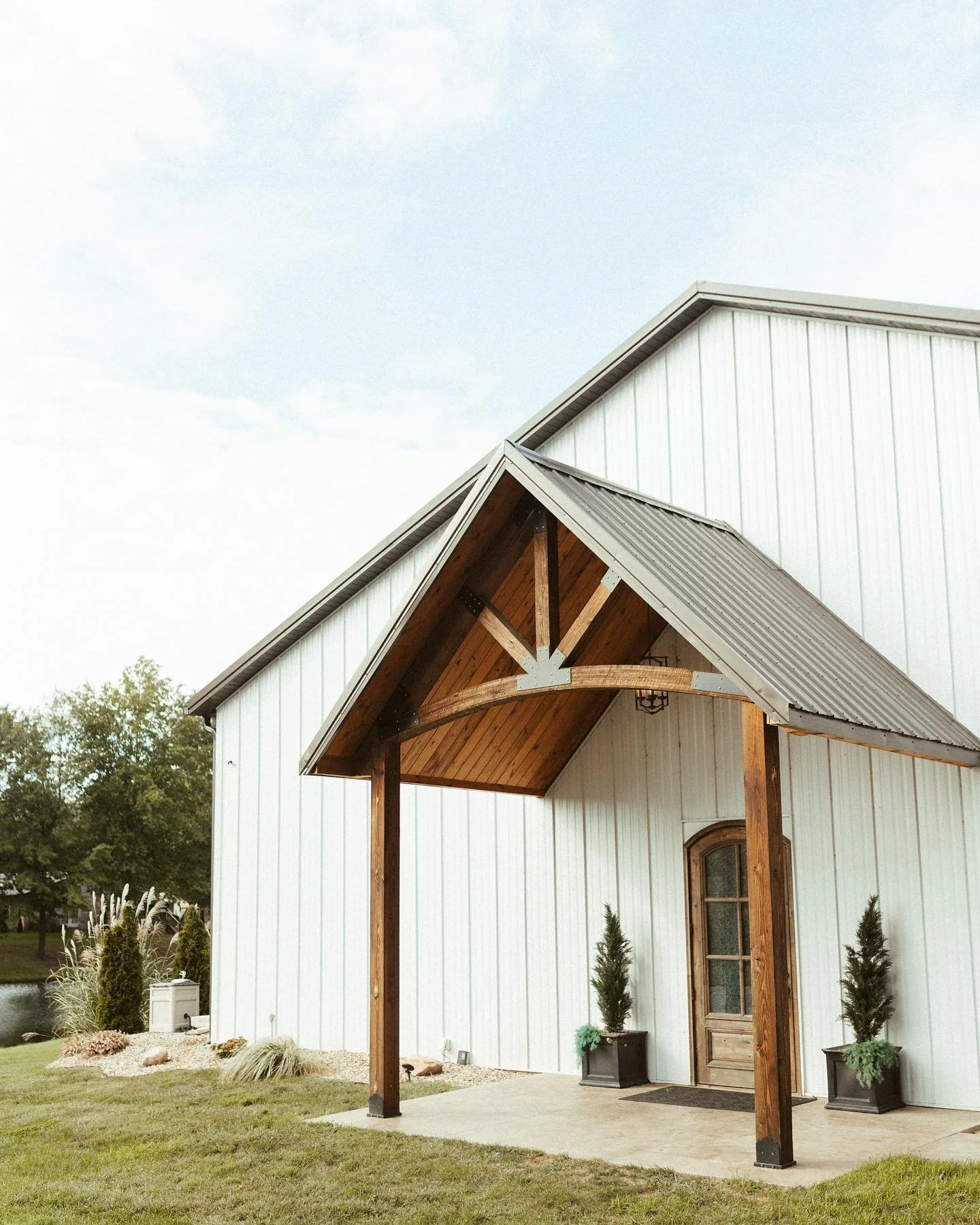 White barn with wooden porch and decorative potted plants outside, surrounded by grass and trees, under a partly cloudy sky.