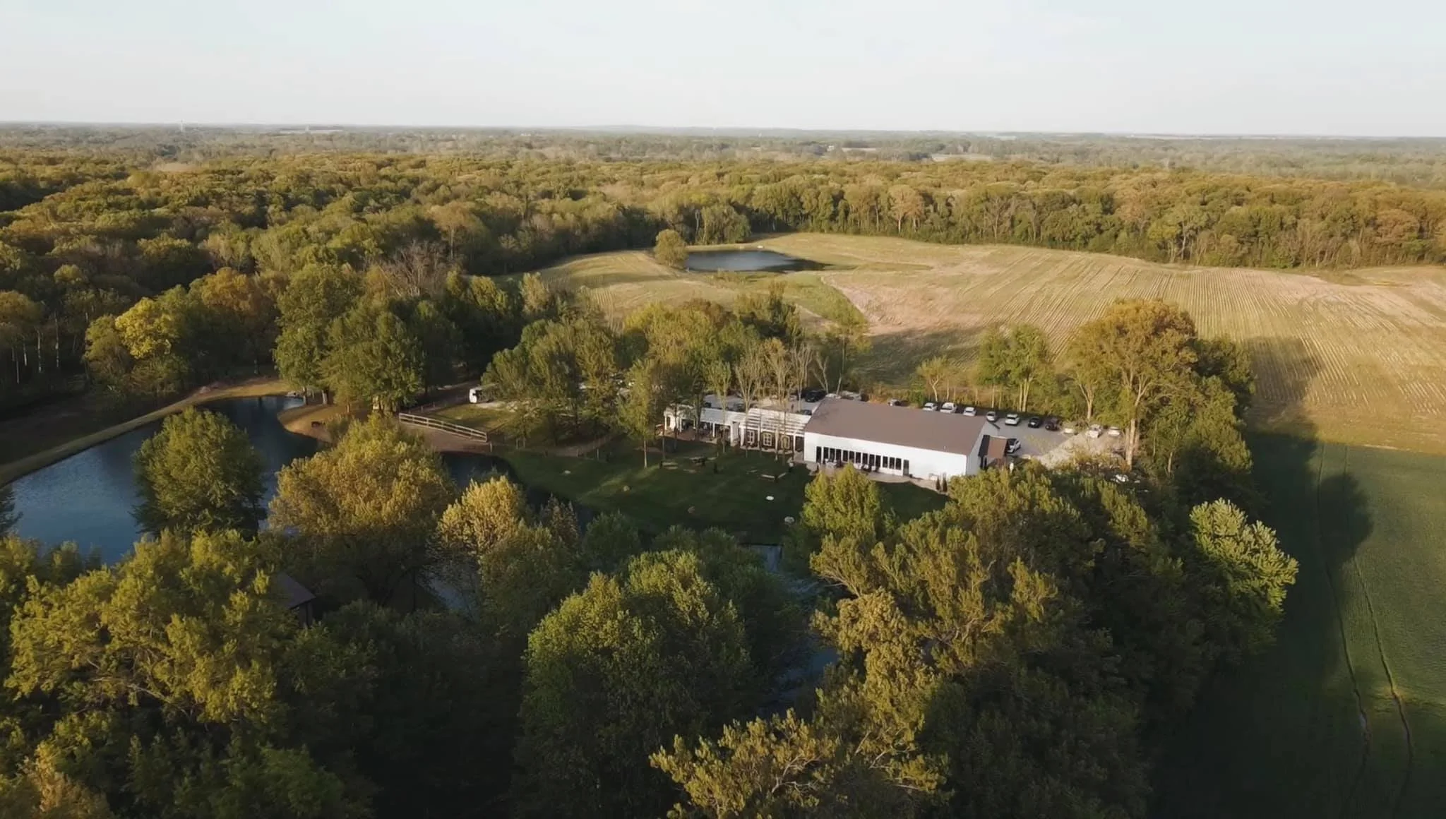 Aerial view of a rural landscape with a white building, parking lot, small pond, trees, and open fields stretching into the distance.