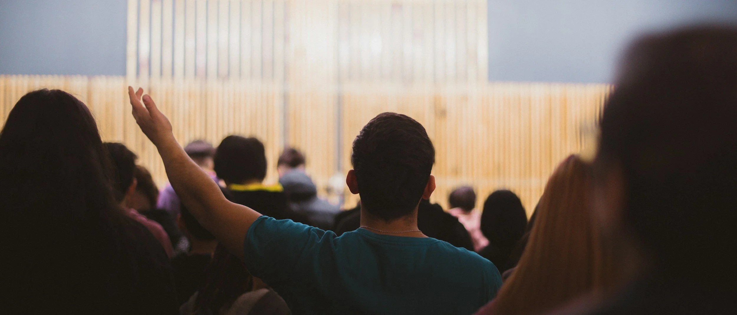 Audience attending a conference or lecture, with a person raising their hand in the middle and a large screen at the front of the room.