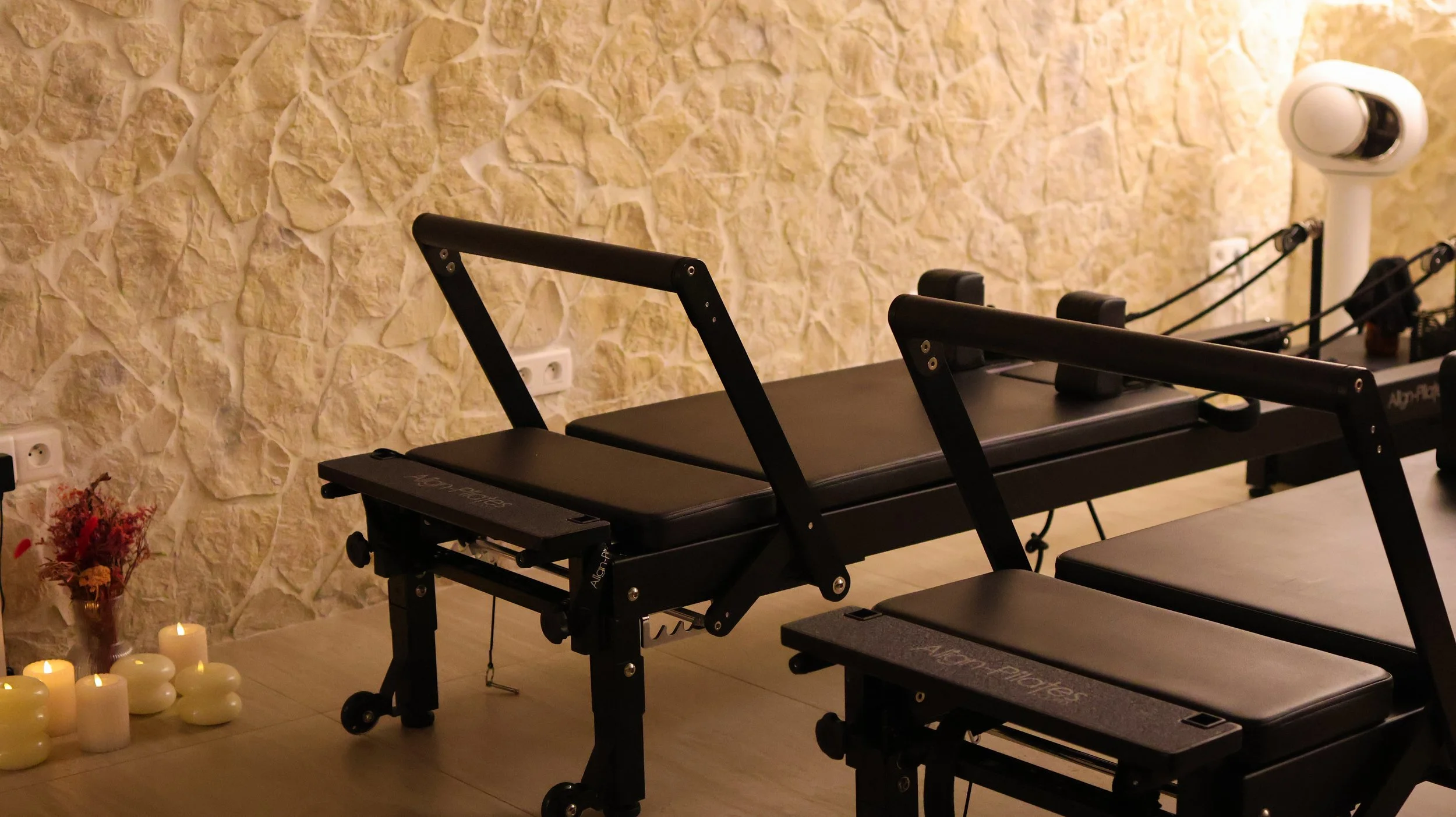 Salle de Pilates équipée de reformers noirs avec un mur en pierre en arrière-plan, ambiance chaleureuse avec bougies et fleurs