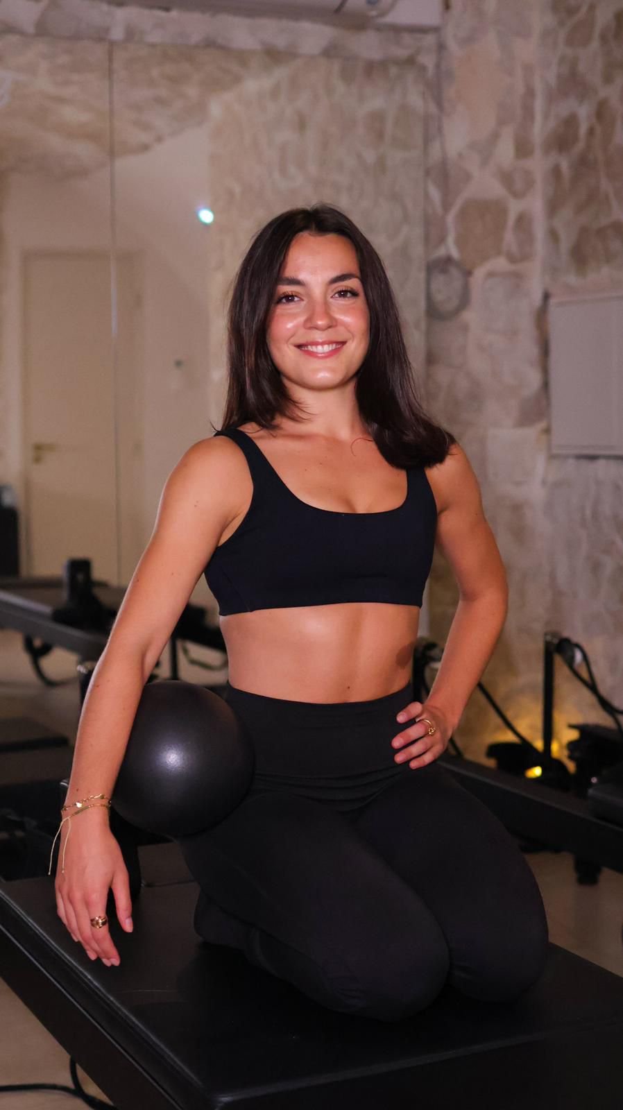 Femme souriante en tenue de sport assise sur un tapis, avec un ballon de médecine à côté, dans une salle de sport avec murs en pierre.