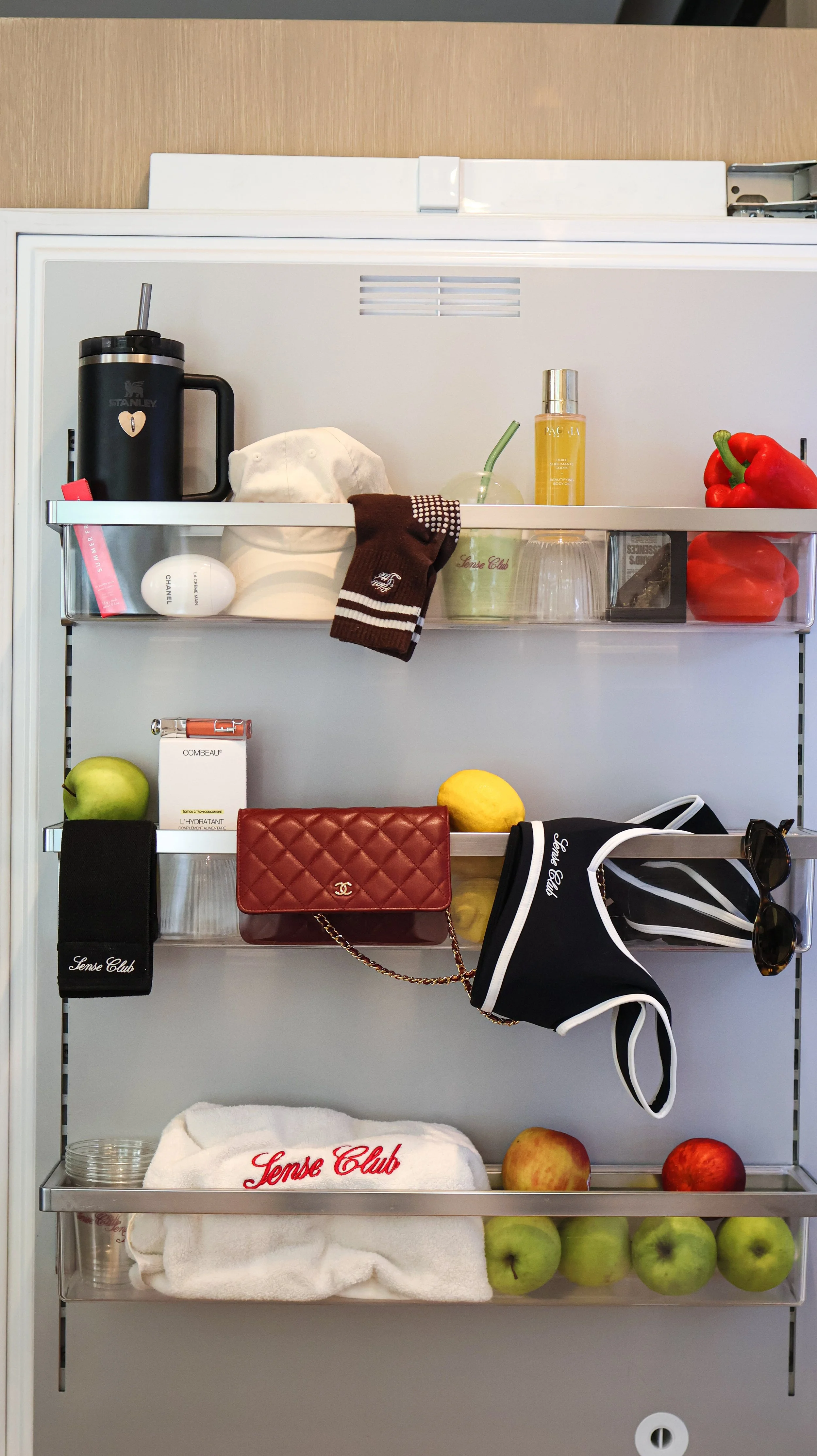 Étagère murale avec divers objets, fruits et accessoires de mode, incluant des pommes, un sac à main, lunettes de soleil, crème et serviette, dans un intérieur moderne.