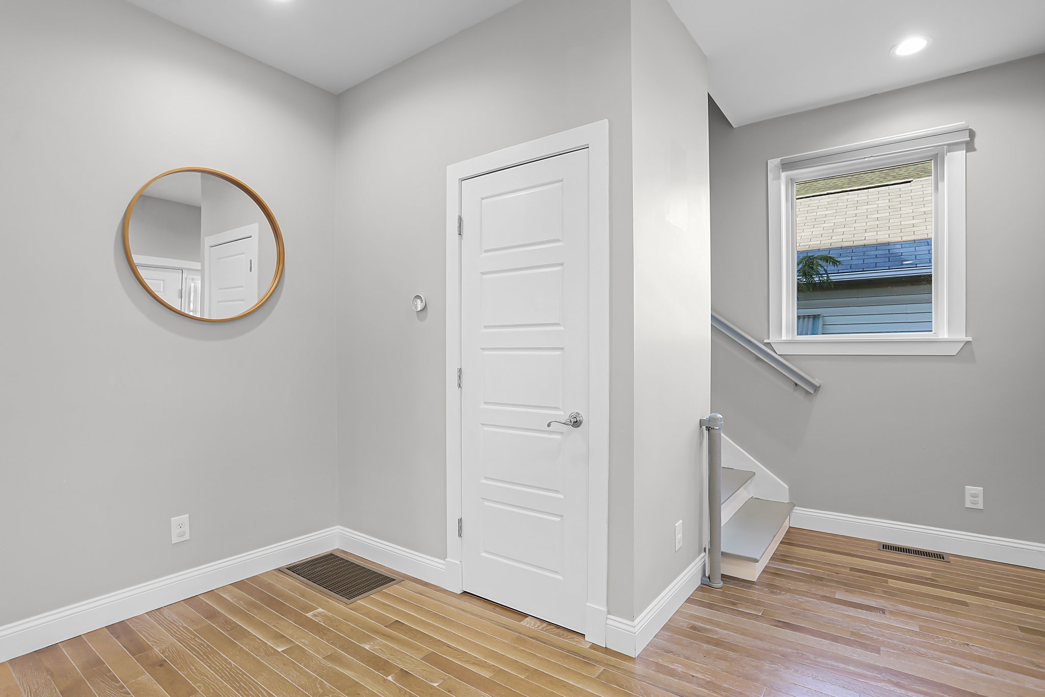 A bright, modern foyer area with a white door, a circular mirror with a wooden frame on the wall, hardwood flooring, a stairway with a white railing, and a window with white trim showing a building with a brick and painted exterior.