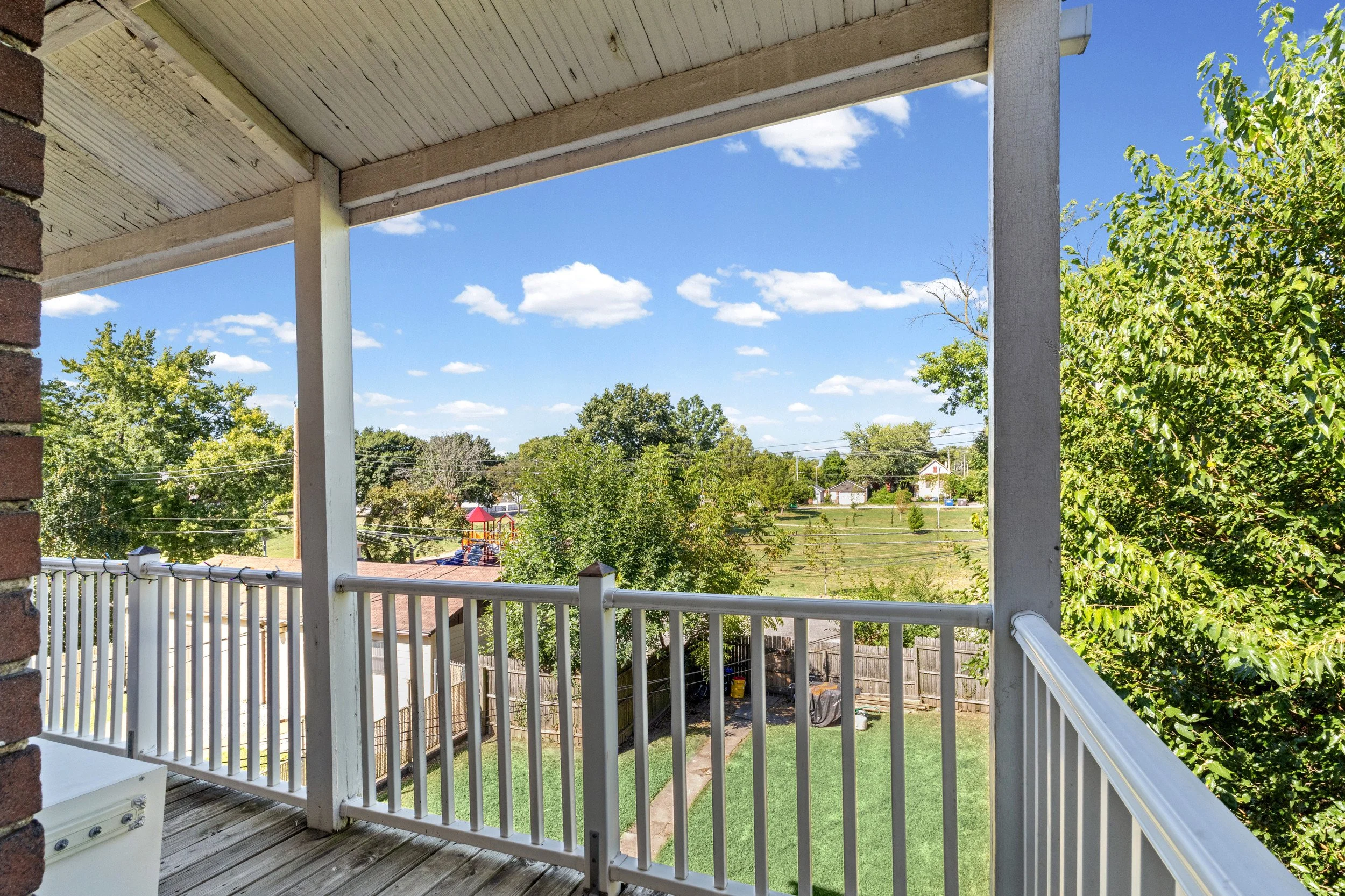 A view from a porch or balcony overlooking a backyard with trees, houses, and a playground under a blue sky with scattered clouds.